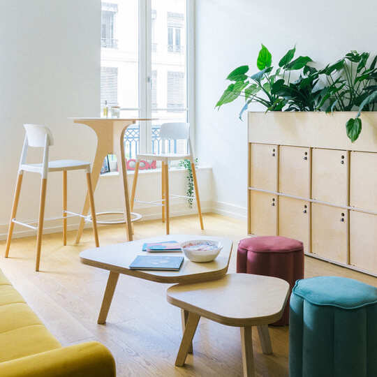 Espace informel au bureau : table haute et basse en bois, poufs, tabourets, canapé et meuble à casiers avec un bac à plantes.