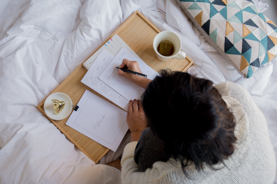 Femme qui écrit, installée confortablement dans son lit, avec une tisane.