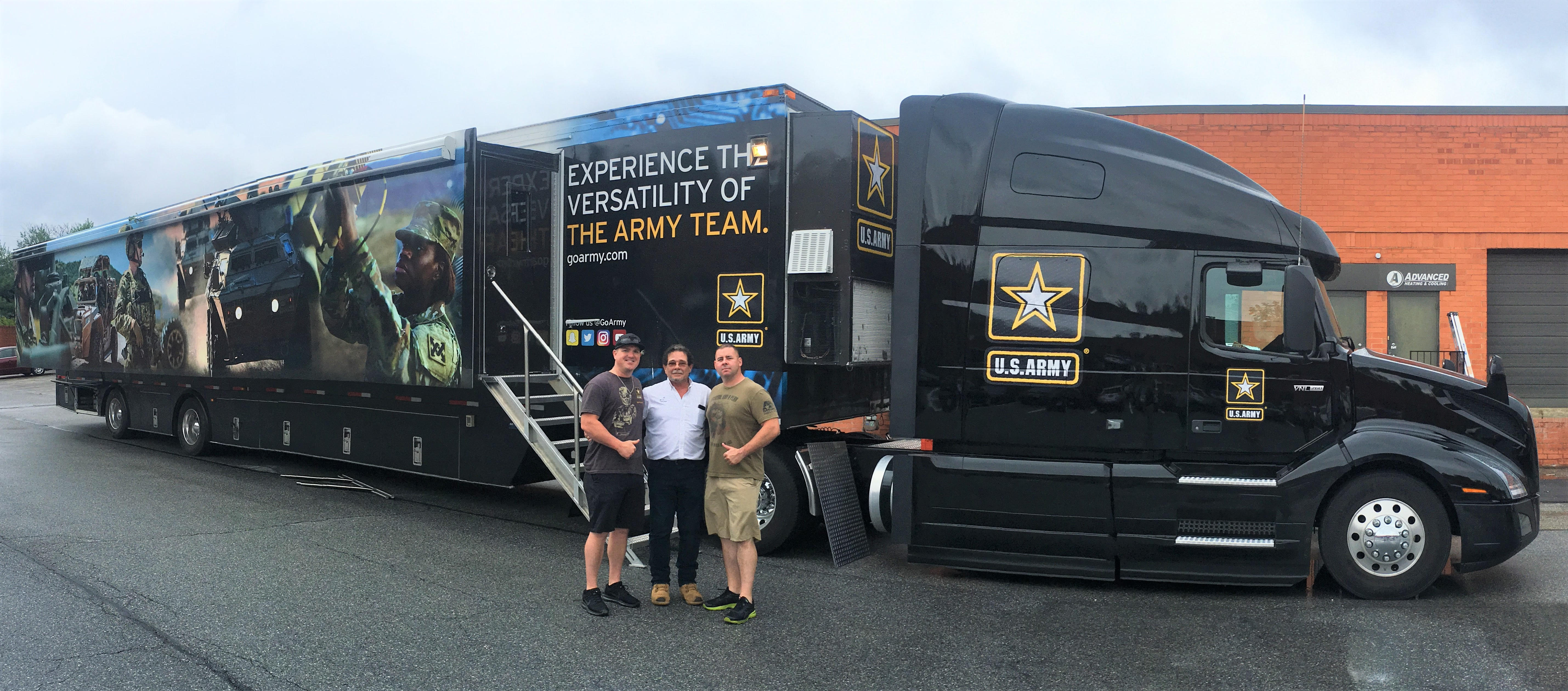 3 men standing and smiling in front of a truck that is advertising for the U.S. Army