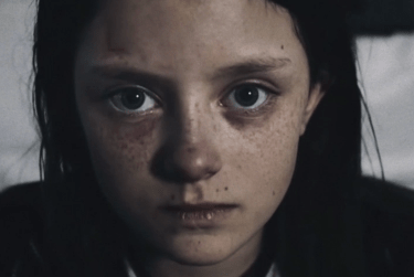 Shot from the a video of a girl with dark hair and freckles staring straight at the camera.
