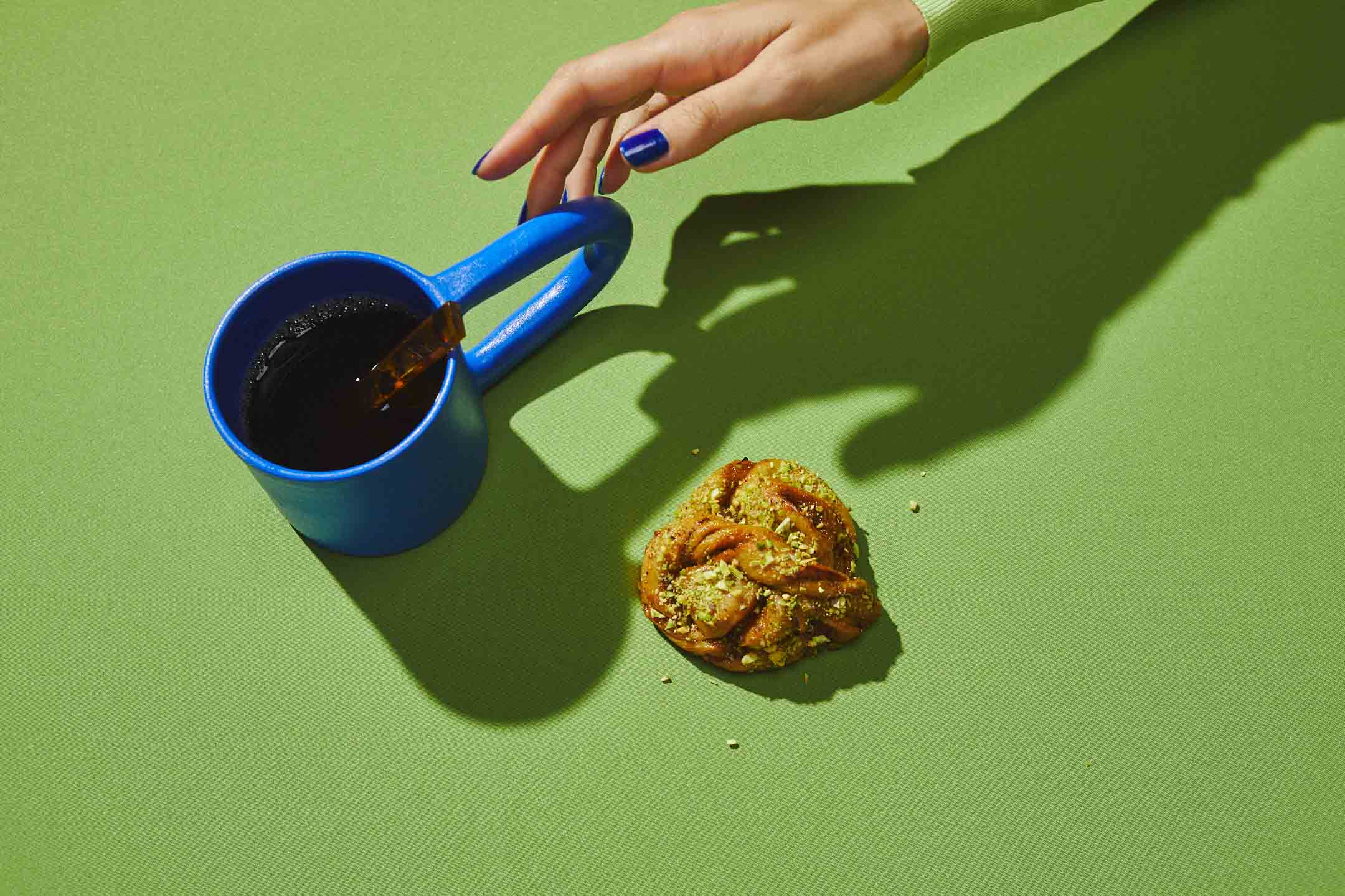 hand reaching for a cup of coffee next to a pastry on a bright lime green background.