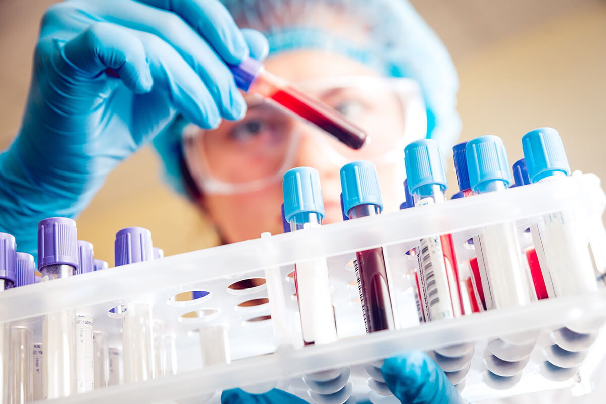 woman wearing lab goggles, latex gloves, and safety cap carrying a vial of blood and looking at it in a lab