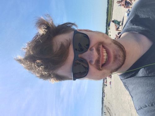 A selfie of Cormac Kenney taken on a beach. His hair is very messed up from the wind, and he looks generally unkempt but happy.