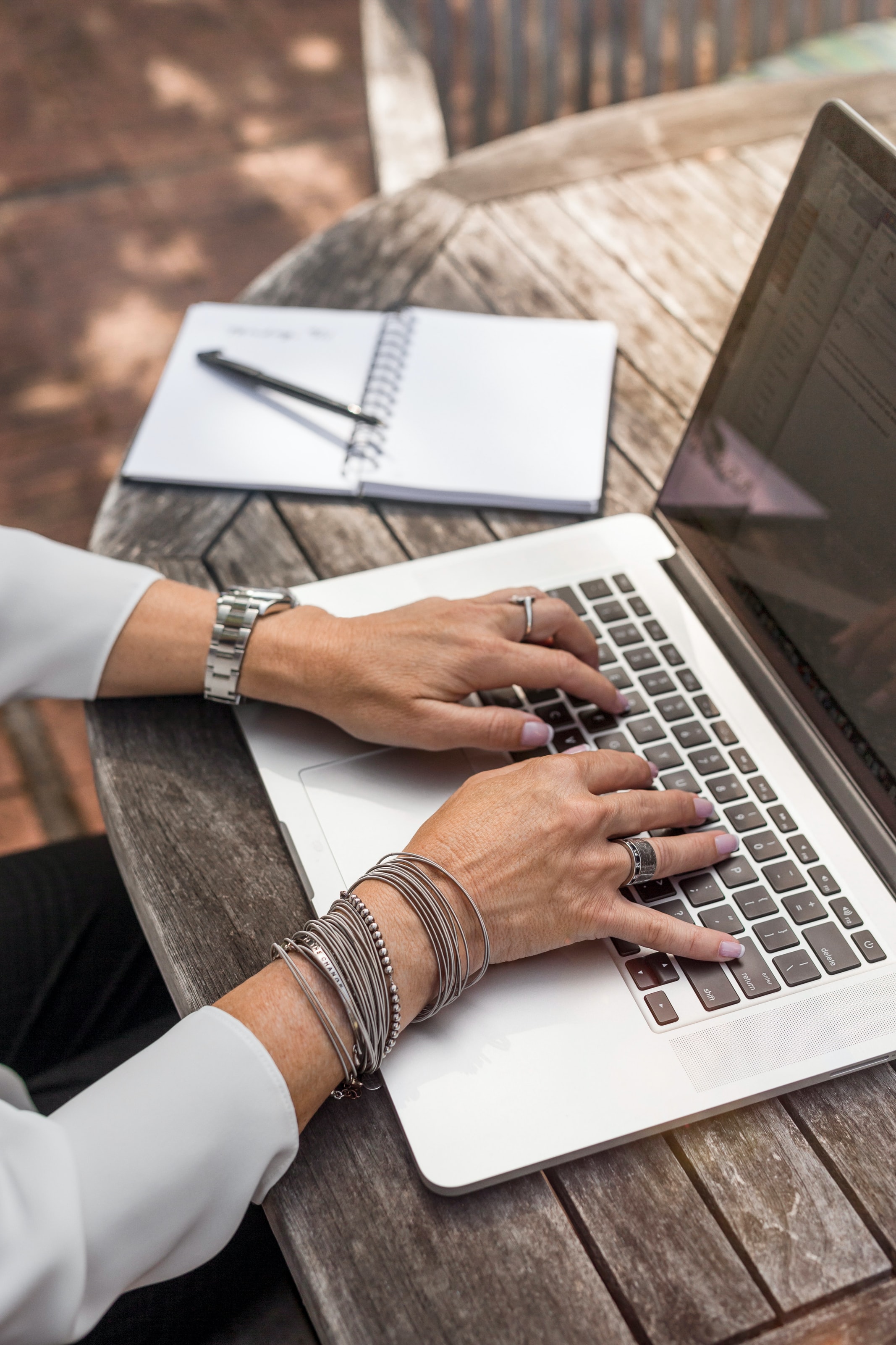 The hands of a person typing on a laptop with a notebook and a pen next to the laptop