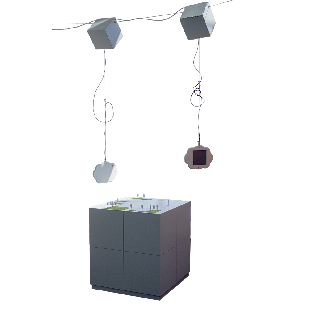 An image showing the complete exhibit. It consists of a white, cube-shaped table standing on the floor and two cloud-shaped devices with displays hanging from a wire rope stretched over the heads of the visitors.