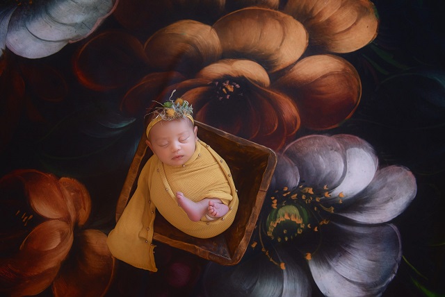 A photograph of a newborn baby swaddled in a yellow blanked lying in a basket with floral background