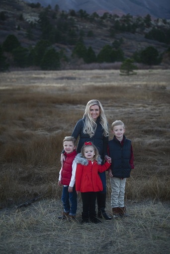 A photograph of a family of four containing a mom and three kids set against nature and mountain background