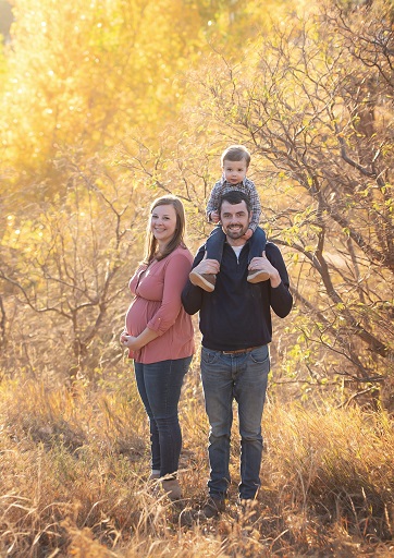 Photograph of family in forest, there is a dad with a son on his shoulders and a pregnant mom holding her belly