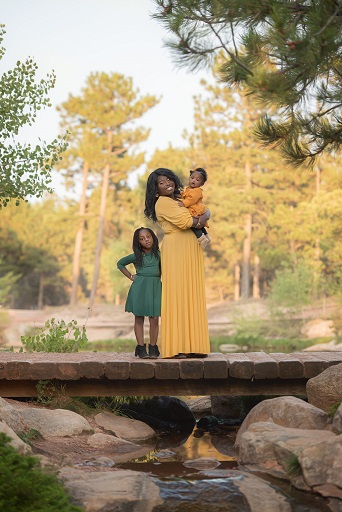 A photograph of a family of three containing a mom and two kids on a bridge with a forest background