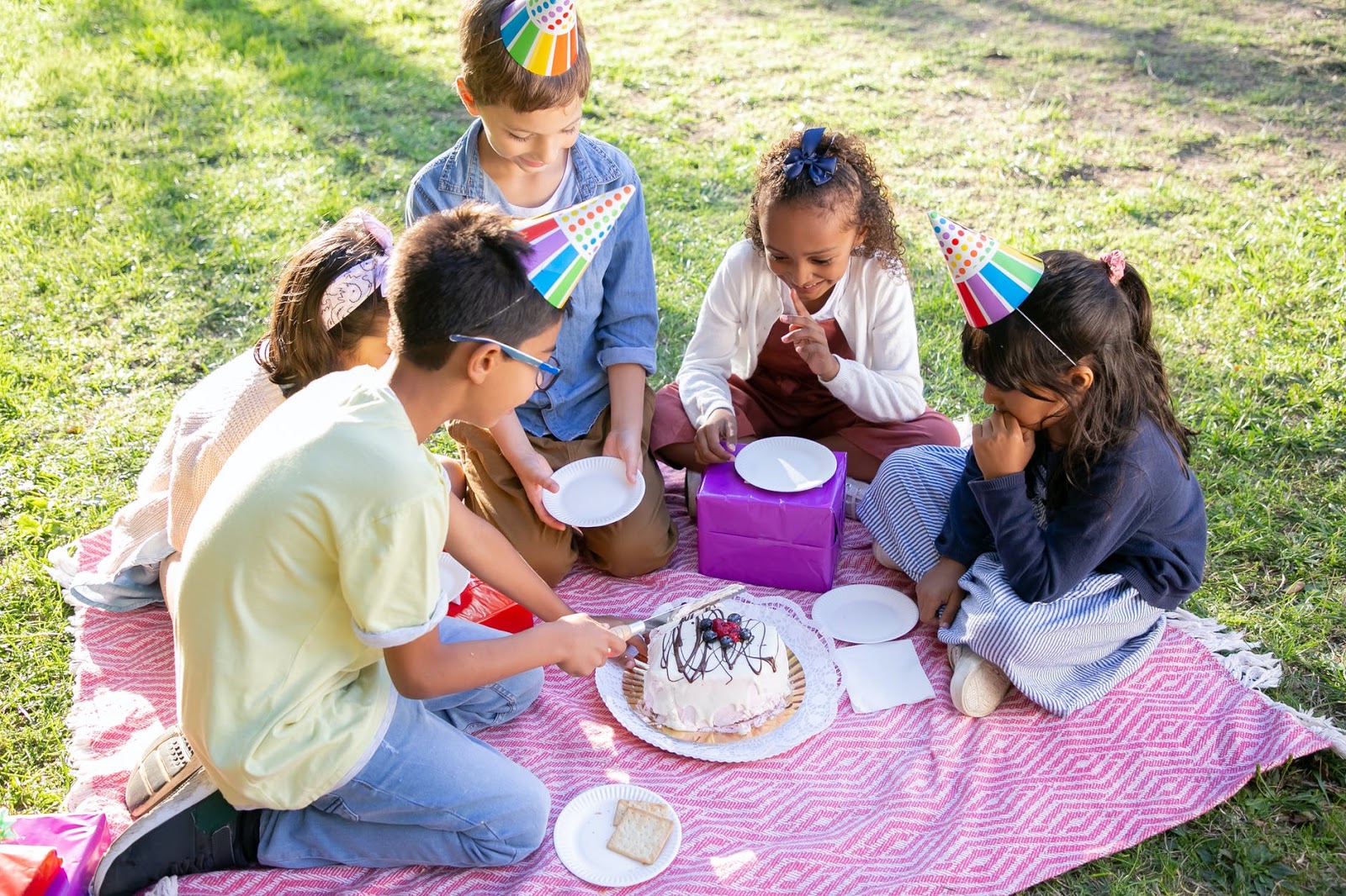 Children sit on a blanket outside and cut into a birthday cake