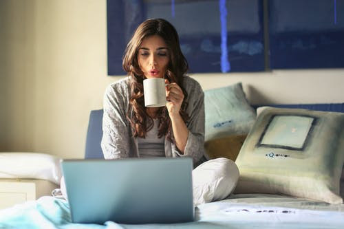 Woman relaxed on bed, drinking from a mug and typing on laptop.