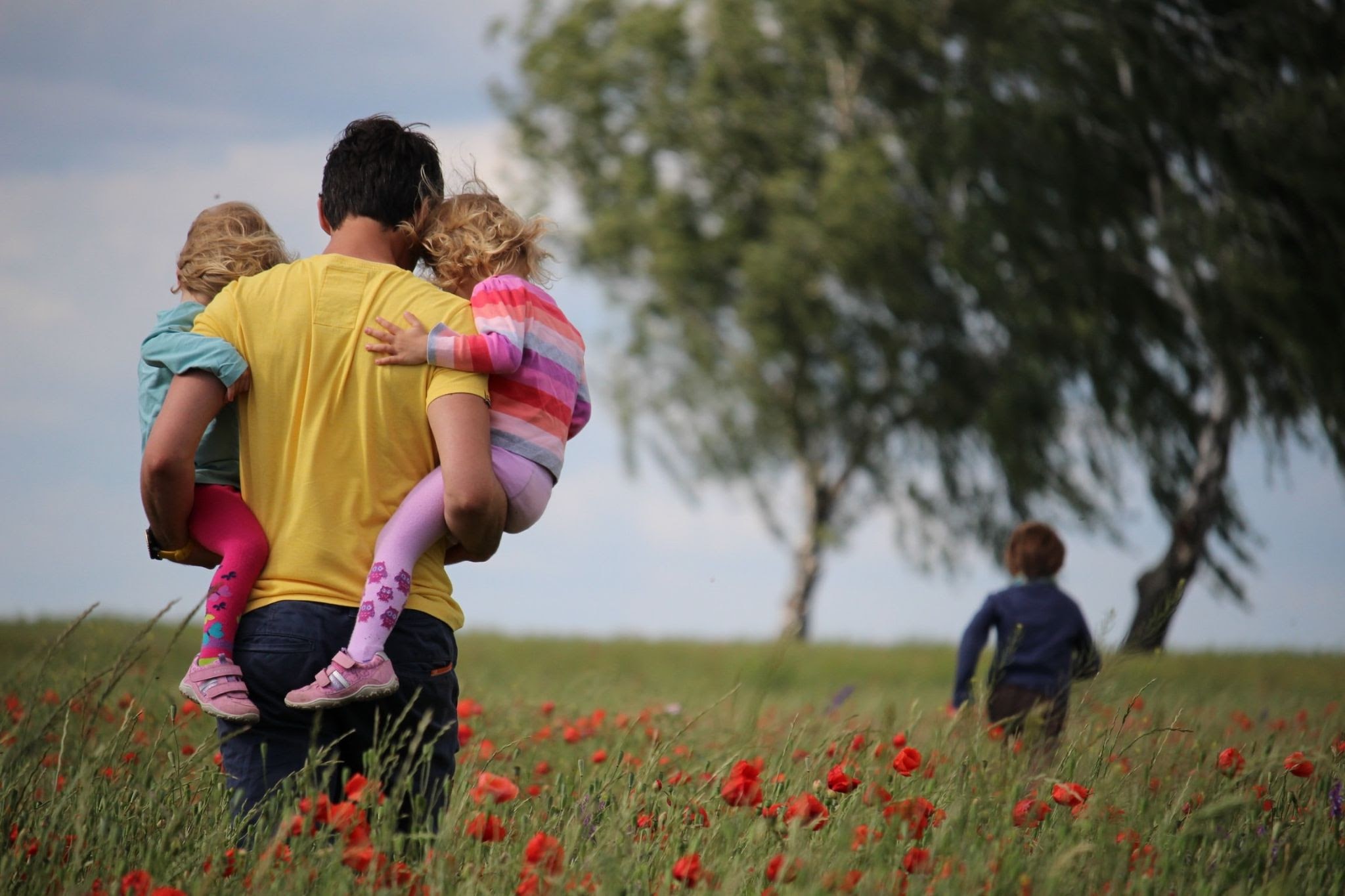 Man holding two children and walking in a field while another child runs ahead