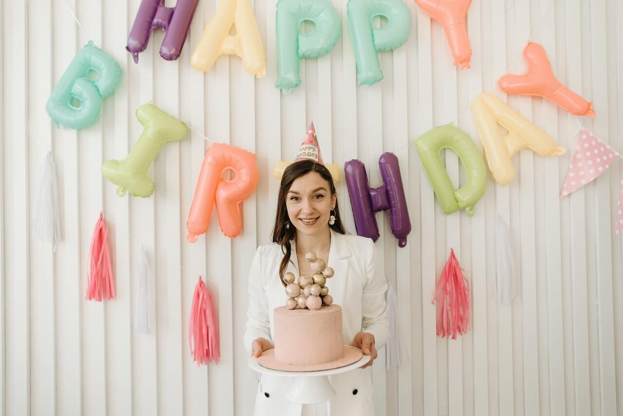 Woman holding a cake, standing in front of a wall with 'Happy Birthday' colorful balloons on it.