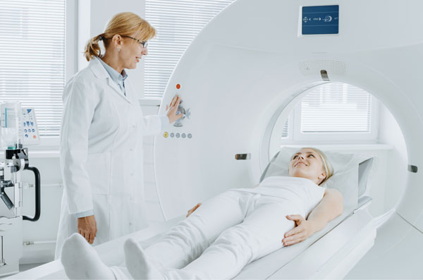 Female doctor is standing over the female patient, the patient lies on the bed of CT machine. Both are dressed white.