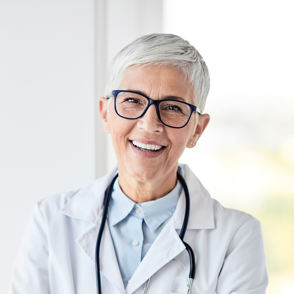 White, smilling woman in white doctor's gown with stetoscopen on her neck.