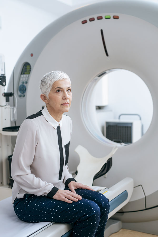 Older woman with short hair sitting on the bed of a tomograph, waiting looking up at somebody.