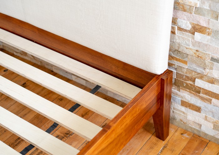 The top right corner of a brown stained bed frame with no mattress and a headboard on a wood floor against a stone wall