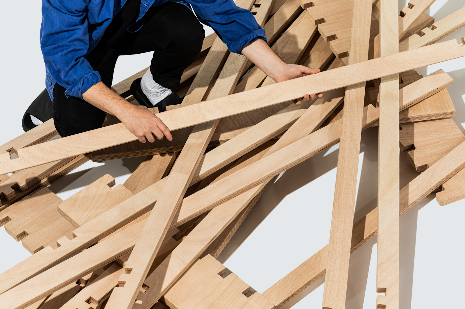 A man with rolled sleeves wearing blue next to a pile of wood slats and holding one in his hands.