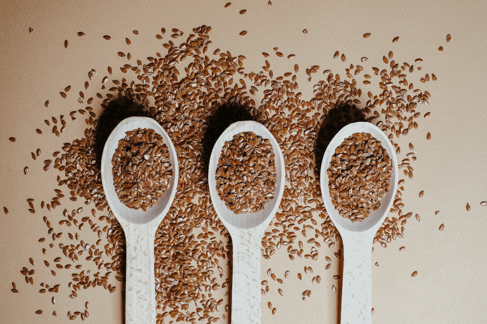Three wooden spoons with flax seeds surrounded by flax seeds on a flat brown surface.