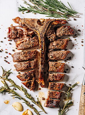 A t-bone beef steak with black pepper, garlic cloves, sprigs of rosemary, and a meat fork on a marble background.