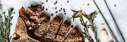 A t-bone beef steak with black pepper, garlic cloves, sprigs of rosemary, and a meat fork on a marble background.