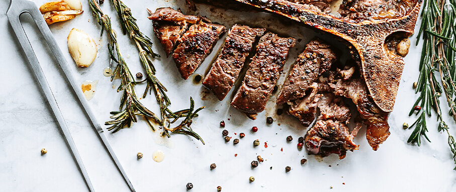 A t-bone beef steak with black pepper, garlic cloves, sprigs of rosemary, and a meat fork on a marble background