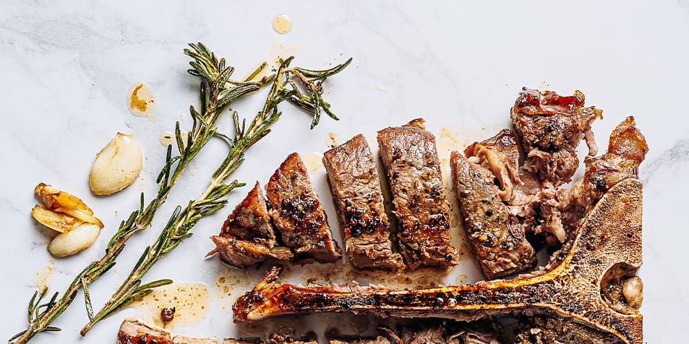 A t-bone beef steak chopped up with garlic cloves and sprigs of rosemary on a marble background