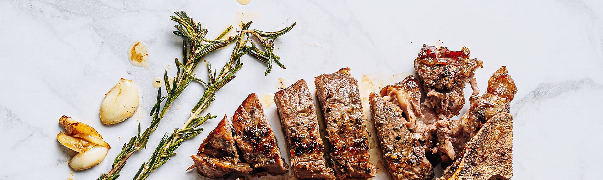A t-bone beef steak chopped up with garlic cloves and sprigs of rosemary on a marble background