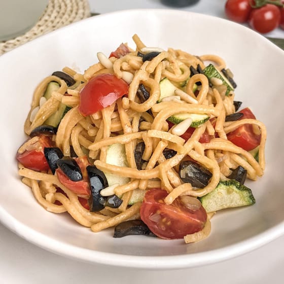 A bowl of lentil spaghetti seasoned with zucchini, black olives, cherry tomatoes and pine nuts.