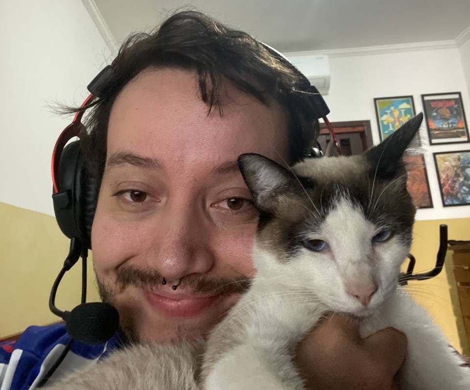 Tassio with a headset on smiles at the camera while lifting cat into frame