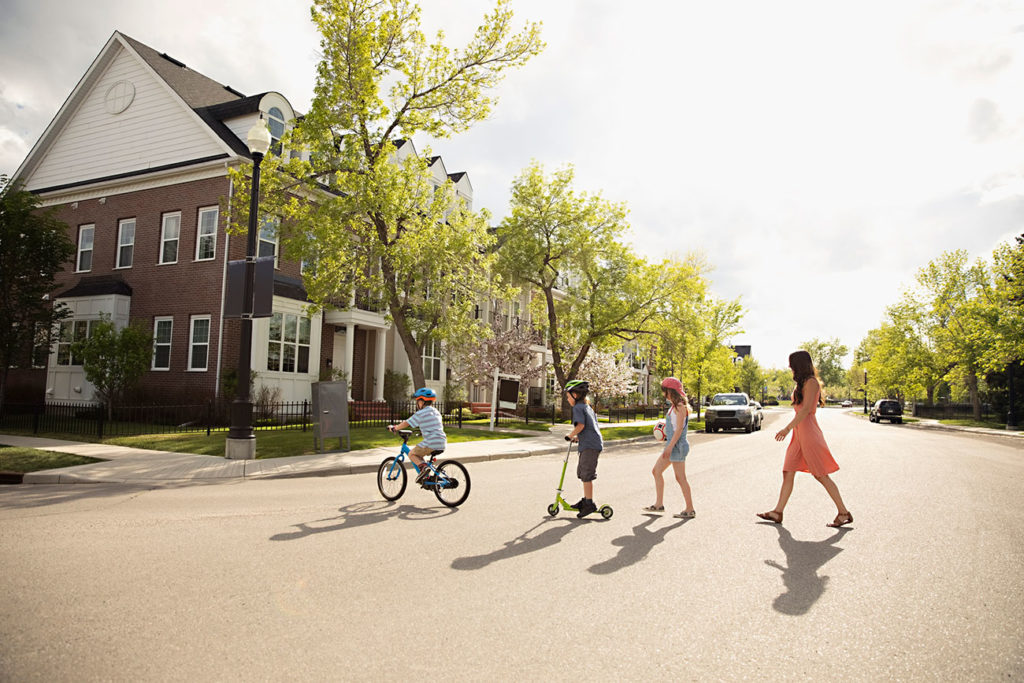 Mother and kids walking and riding a scooter across a street on a sunny day