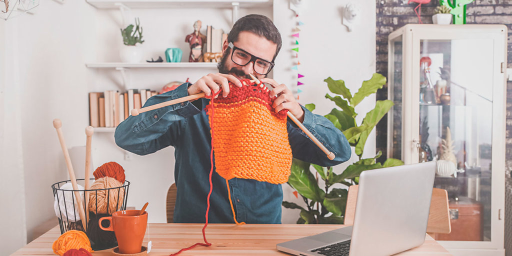 Bearded man knitting at home using laptop for watching online tutorial