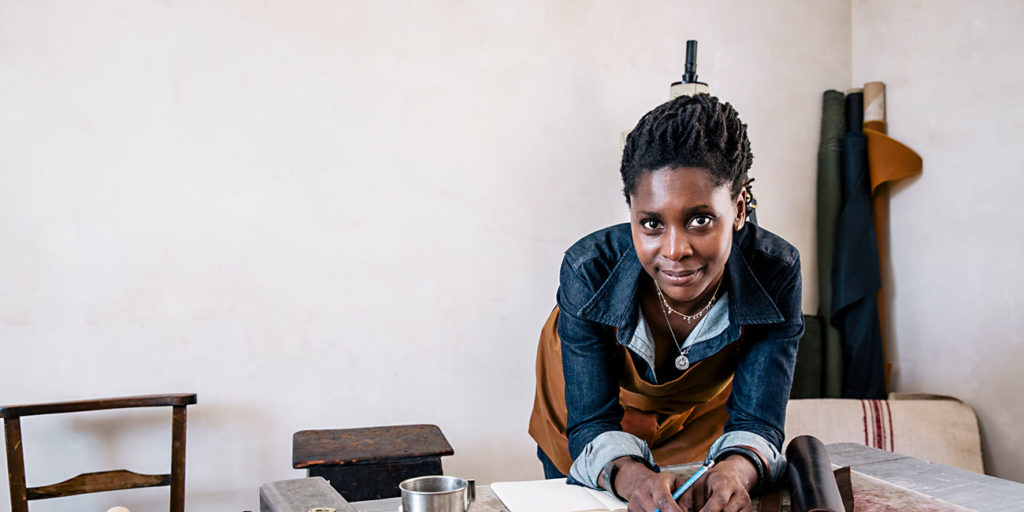 a self-employed craftswoman poses for a portrait, at work designing handbags