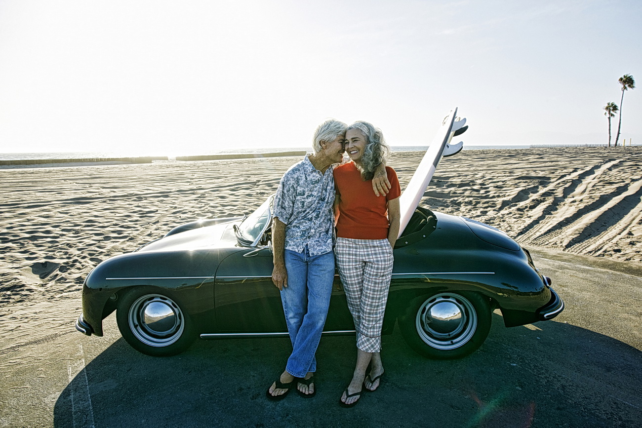 Older Caucasian couple leaning on convertible car with surfboard on beach