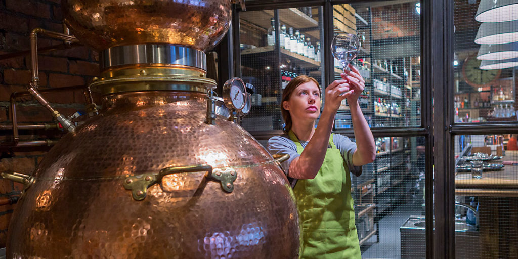 A female worker is sampling the alcohol produced from the copper distillery boiler used to produce gin