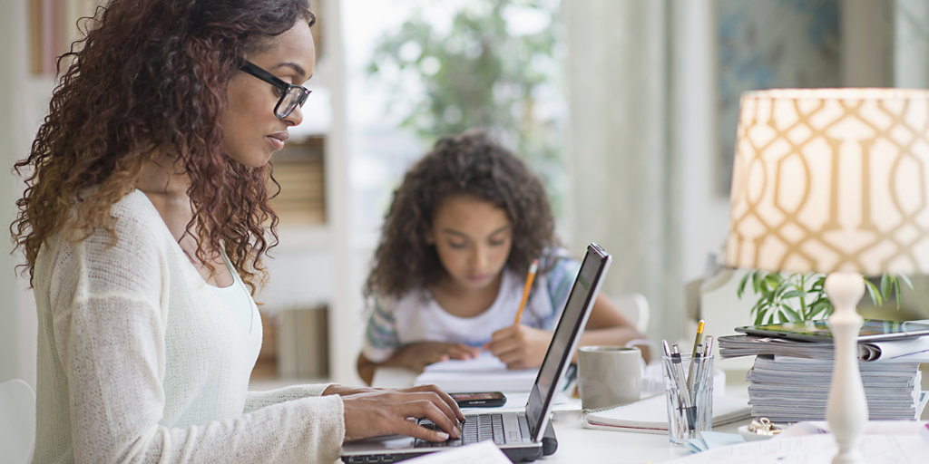 Woman working home using laptop, daughter doing homework in background