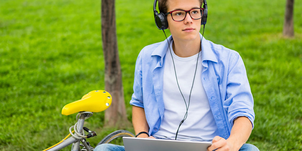 Portrait of male college student with road bicycle sitting on a bench using laptop and headphones