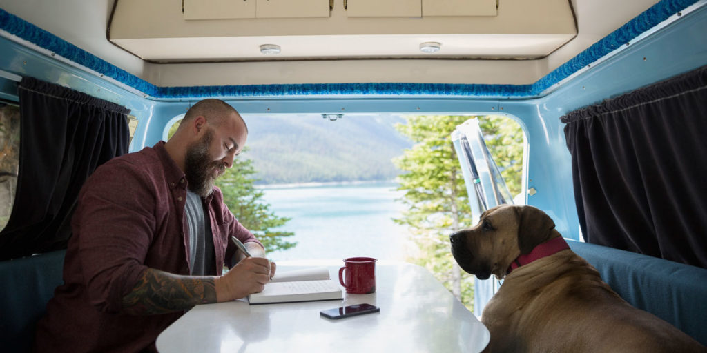 Man with dog writing in journal at table in the back of camper van at lakeside