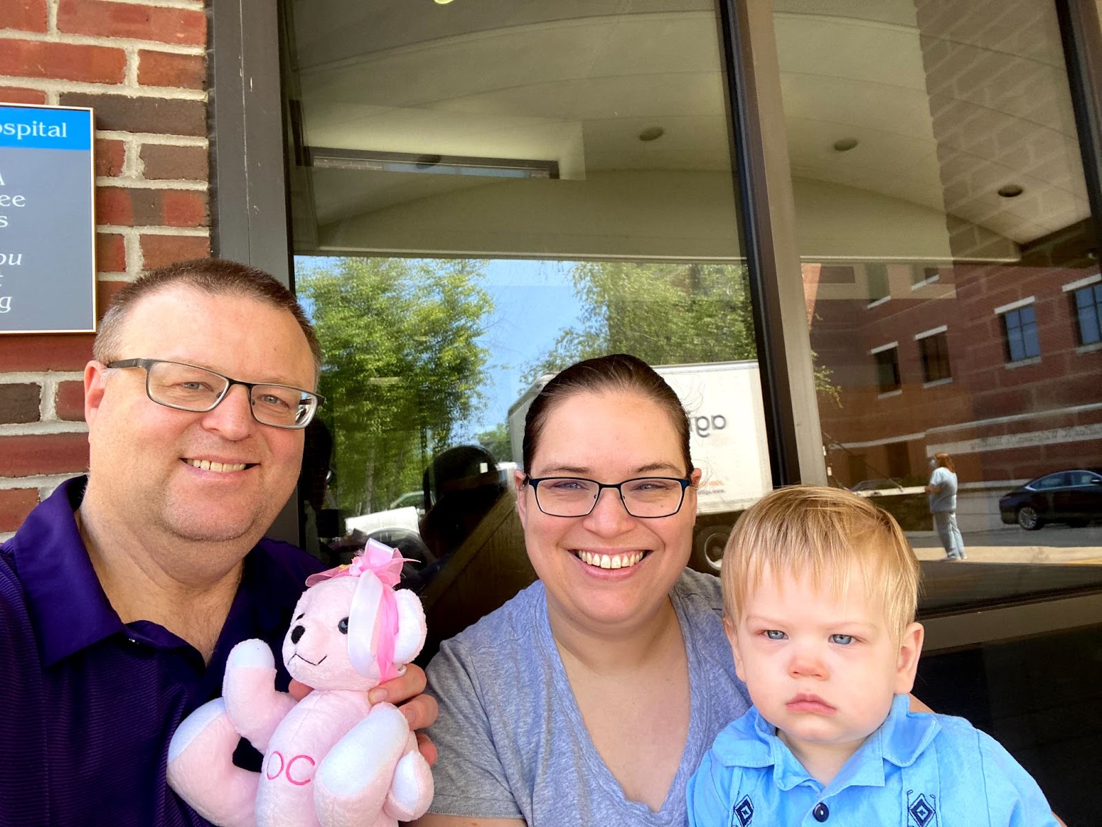 The author of this article and NICU loss Mom, Michelle, sitting in front of a hospital with her husband, her rainbow baby and son, Elliott Miguel, and a pink teddy bear with a pink and white bow that represents her angel baby and daughter Colette Louise. Michelle and her husband are smiling.