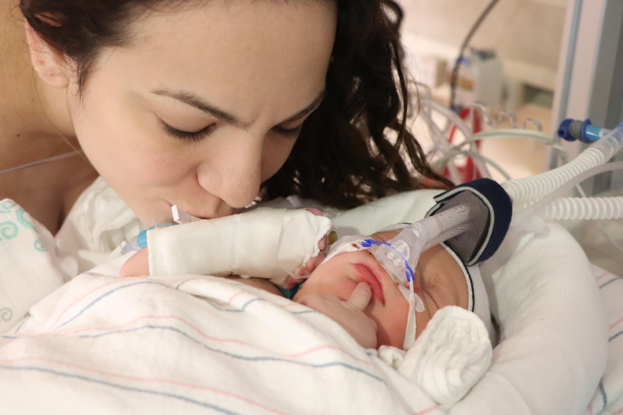 NICU Mom Erica, leaning down with her eyes closed to kiss her NICU Baby, Caden. Caden has his arms wrapped with IVs, and has a breathing tube in. He looks to be just born. You can see the love, gratitude and pain in Erica's face.