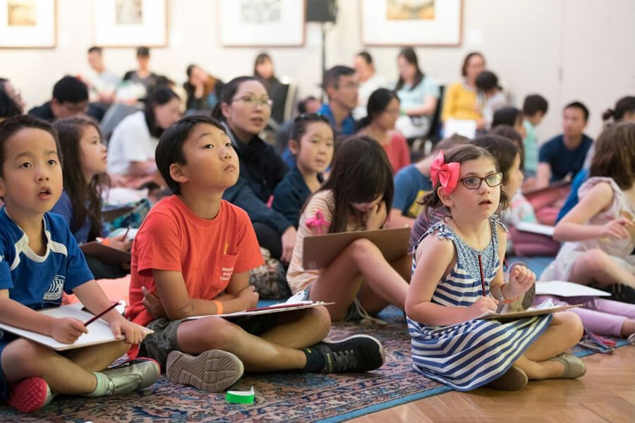 Room full of children sitting cross legged on the floor writing on boards