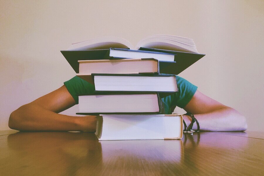 Pile of books on a desk with person behind them with their head on the desk