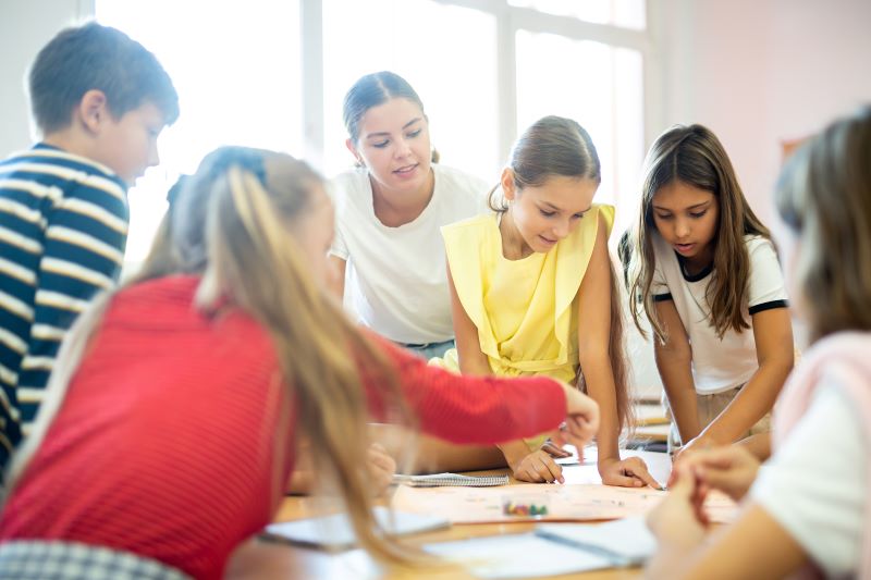 Reforço escolar: conheça 7 estratégias eficientes e inovadoras 1 Foto de um grupo de crianças jogando jogo educacional em cima de uma mesa, acompanhados por uma professora.