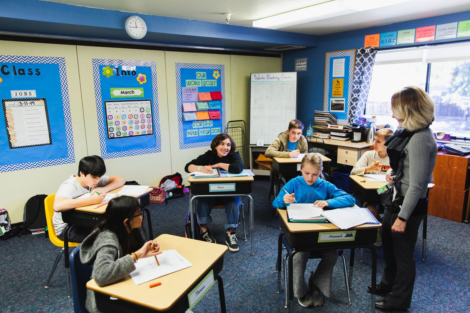 A photo of children sitting in desks and a teacher talking to the students during Language Arts Class.
