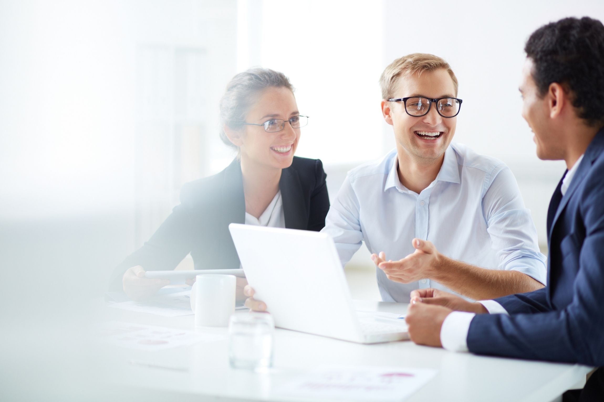 Three people in business attire smiling and chatting at a desk with a laptop.
