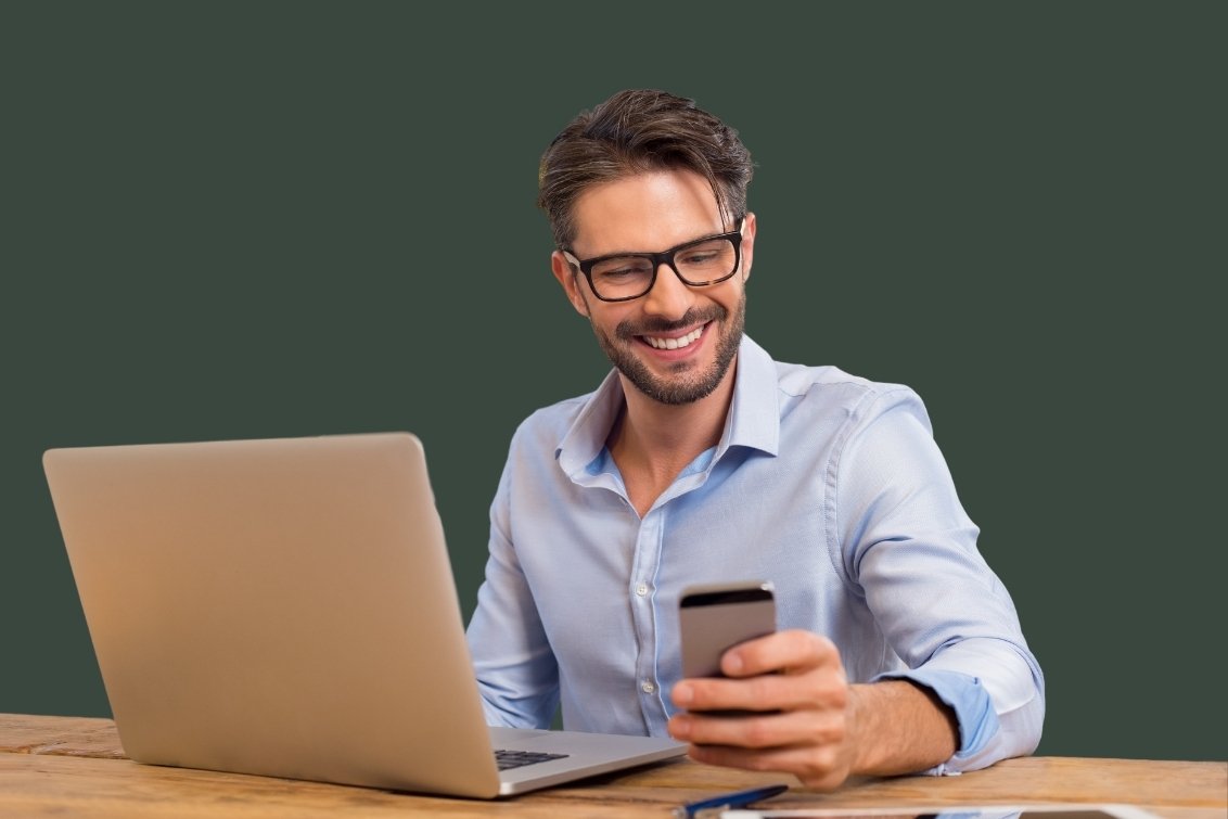 Office worker sitting at desk with laptop and smiling at iPhone.