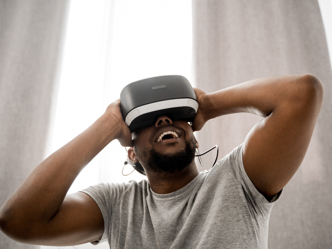A masculine presenting Black person wearing a grey t-shirt VR headset looking upwards.