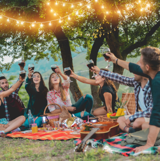 Happy friends having fun at picnic dinner with vintage lights outdoor next vineyard
