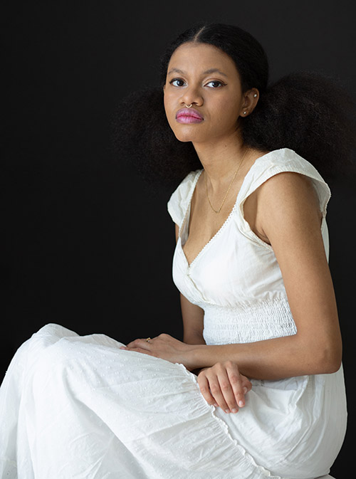A photo of Jaime. They are sitting in a white dress with their arms crossed over their lap, looking at the camera.