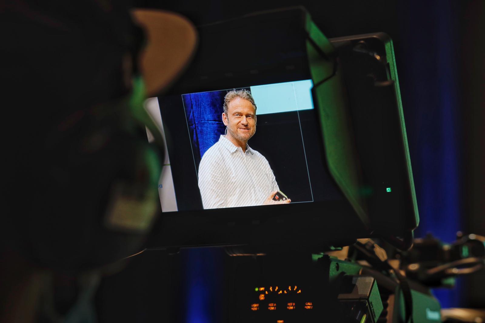 Julian Kawohl is filmed on a stage. A cameraman is seen in the foreground.